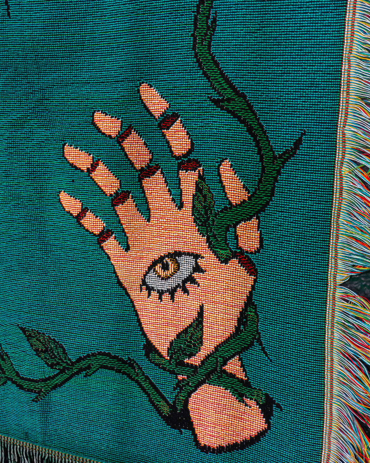 Embroidered design of a hand with an eye and green leaves on a teal fabric.