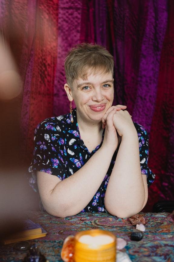 EdenSparkle with short hair and a colorful shirt, sitting at a table with a patterned tablecloth, smiling gently, ready to do your next psychic reading.