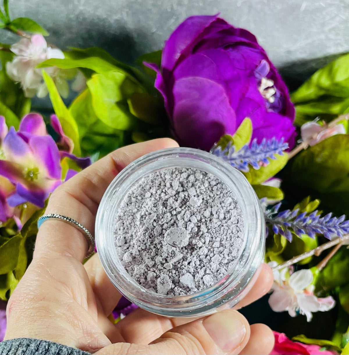 Hand holding a small container of gray powder with flowers in the background