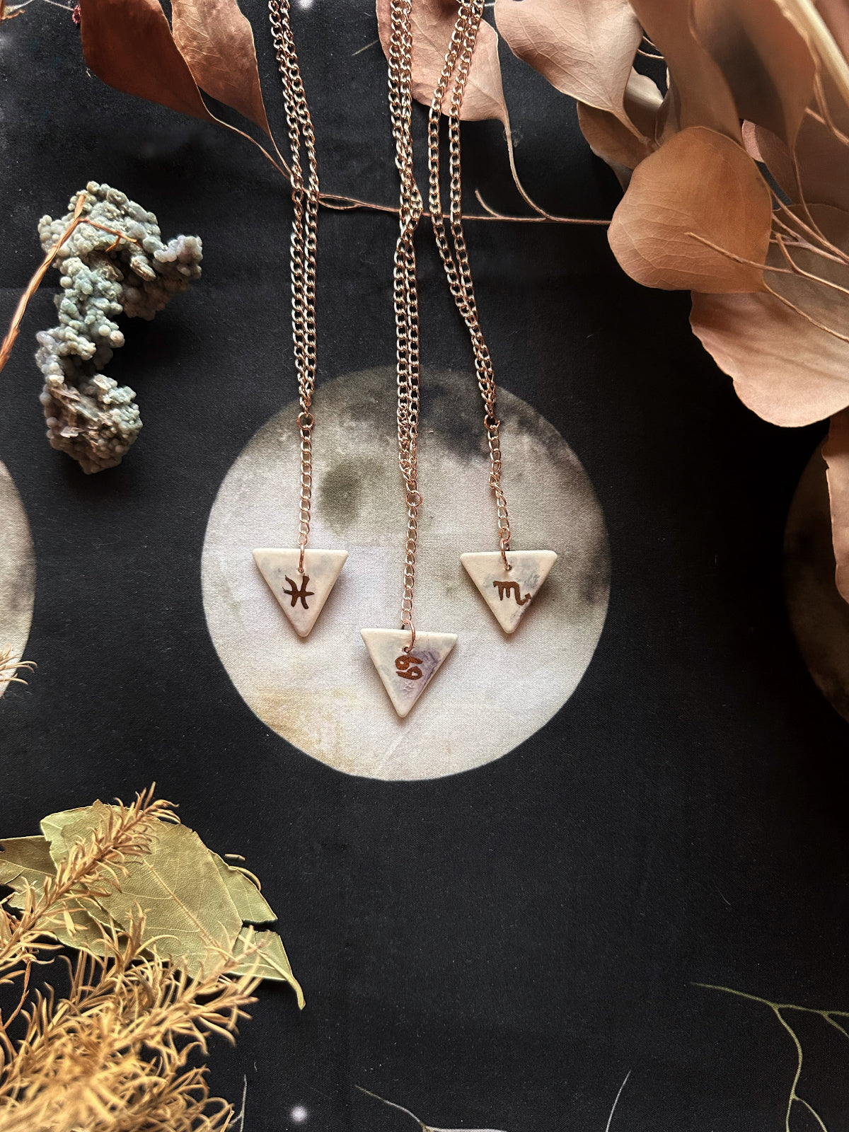 A double-sided porcelain necklace with one side featuring a geometric design and the other with a gold-colored, watercolored underglaze design representing the zodiac sign. The necklace is displayed on a textured background with decorative leaves and a moon.