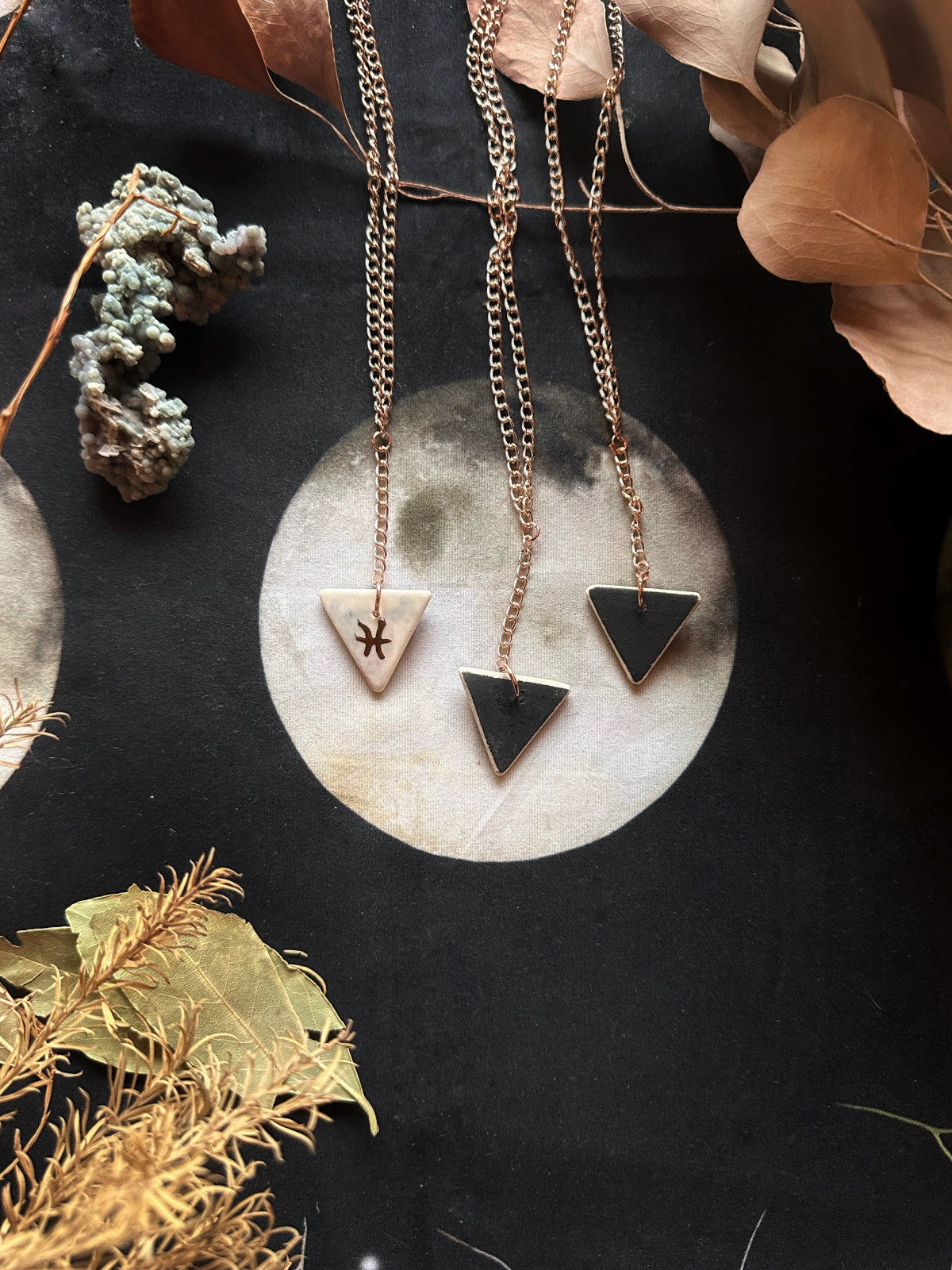 A double-sided porcelain necklace with one side featuring a geometric design and the other side a stylized triangle with a gold outline, displayed on a dark background with natural elements.
