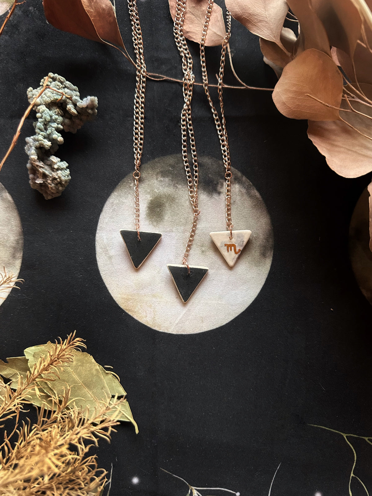 A double-sided porcelain necklace with one side featuring a geometric design and the other with a gold-colored, watercolored underglaze design representing the zodiac sign. The necklace is displayed on a textured background with decorative leaves and a moon.