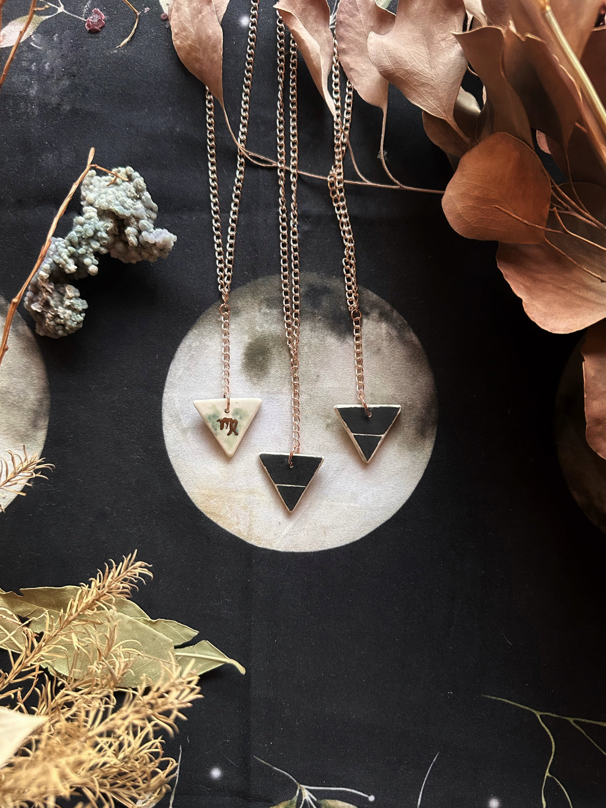 A double-sided porcelain necklace with one side featuring a geometric element design and the other side a gold-colored zodiac sign symbol, displayed against a moonlit background with natural decorations.