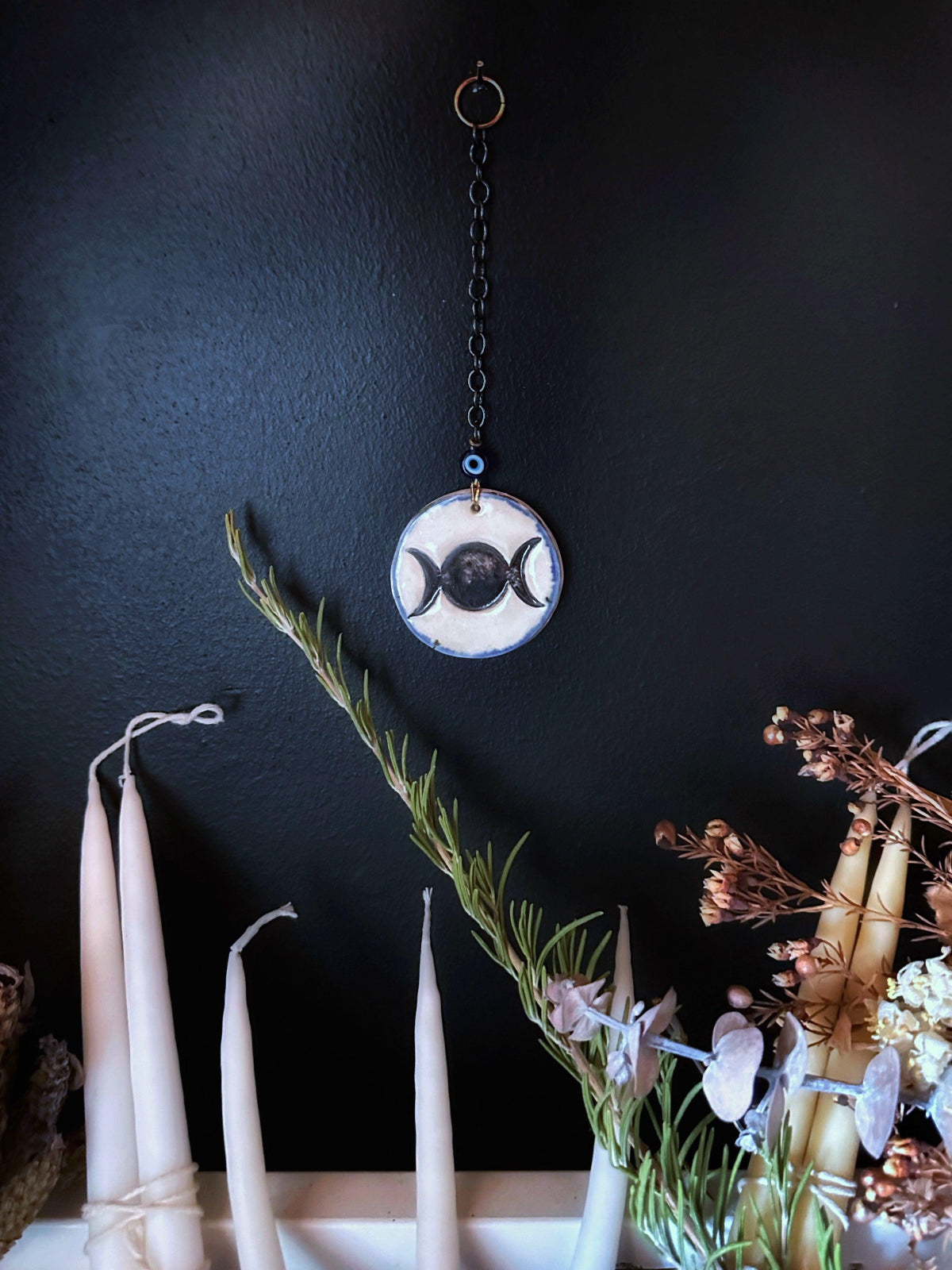 A handcrafted porcelain amulet with a blue halo, Turkish glass bead, and gun metal chain, displayed as a wall ornament with candles and decorative branches in the background.