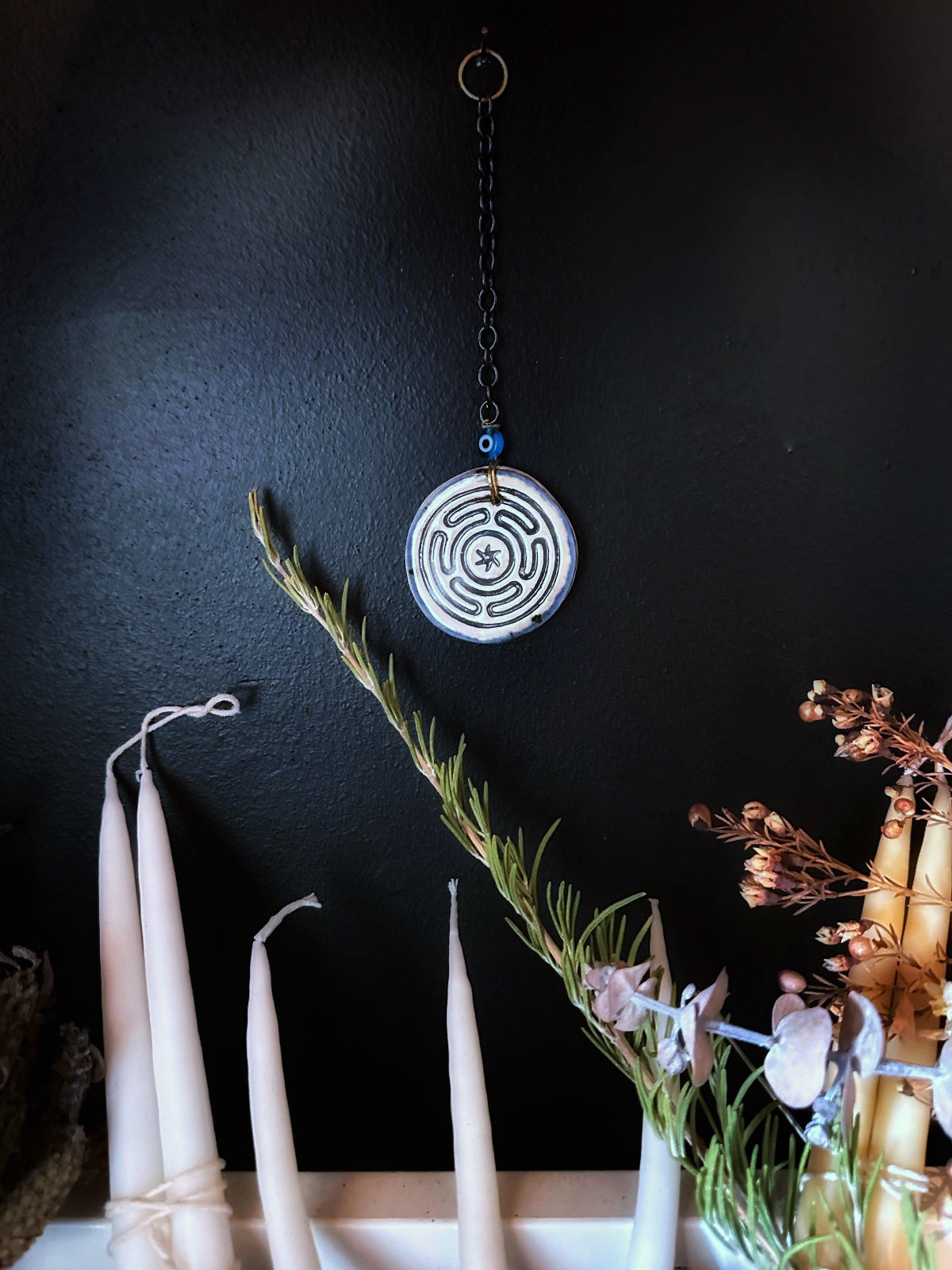A handcrafted porcelain amulet with a geometric pattern, featuring a blue lined sketch pattern on the back, displayed on a dark wall with a black chain and a decorative branch with dried flowers next to it.