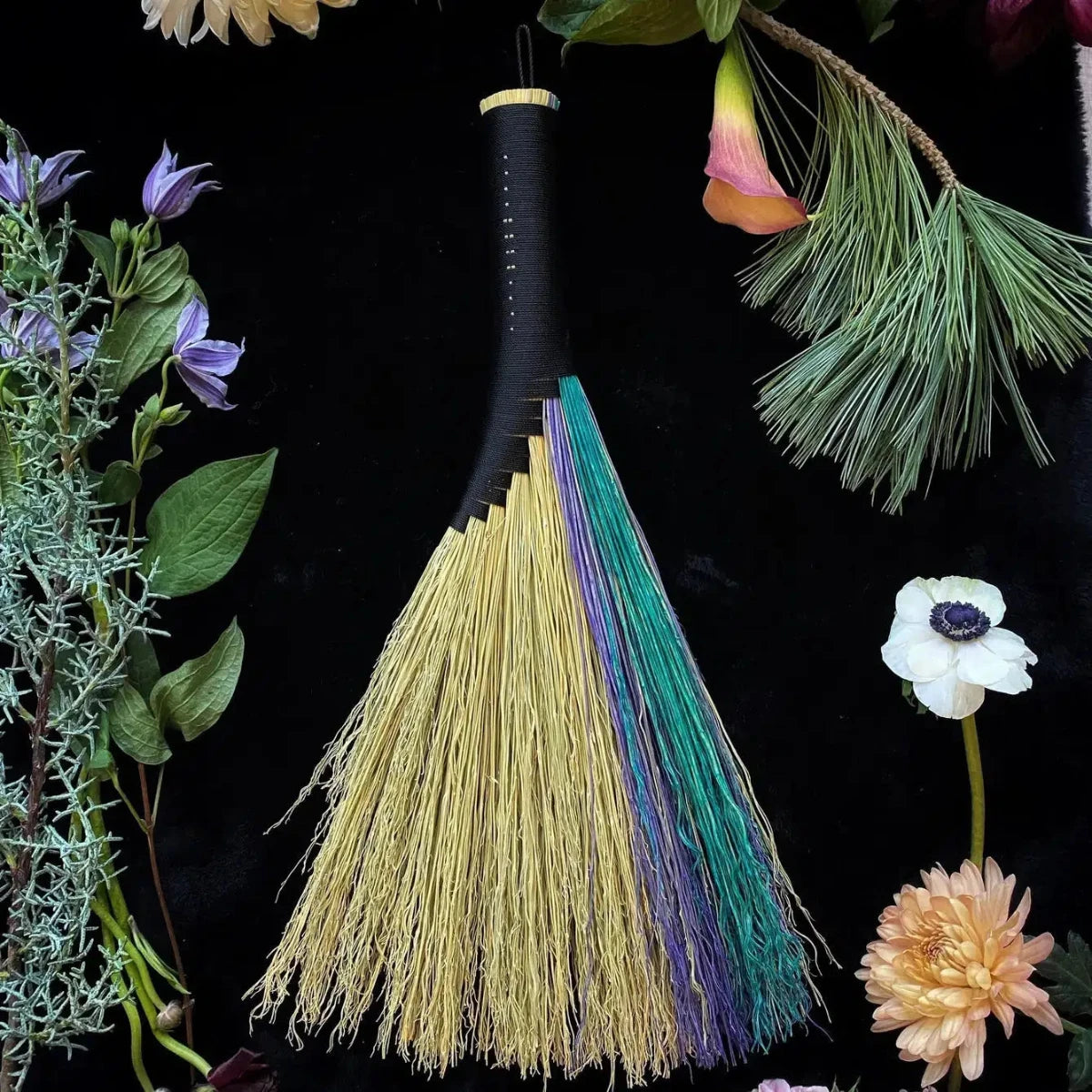 A hand woven black broom with multicolored streaks, displayed with a variety of dried flowers and greenery in the background.