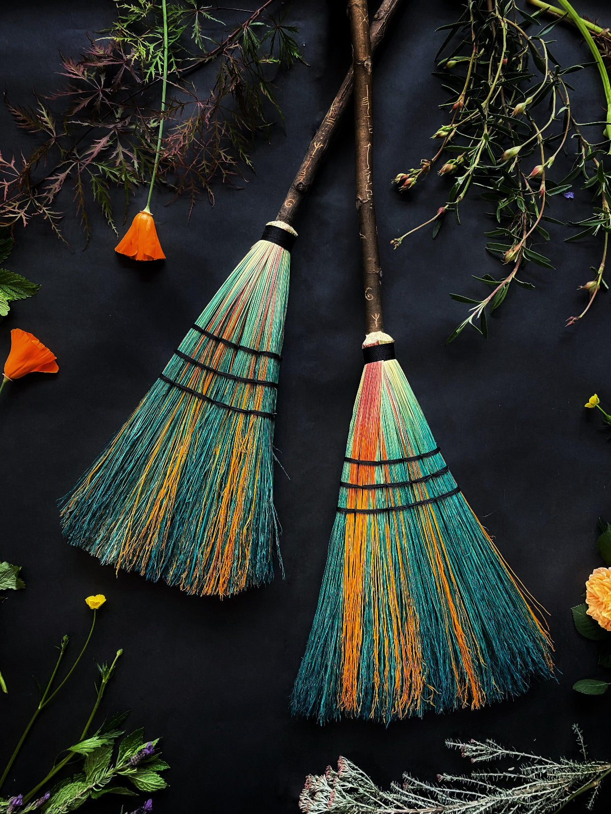 Two colorful brooms with wooden handles and bristles arranged on a dark surface surrounded by herbs and flowers.