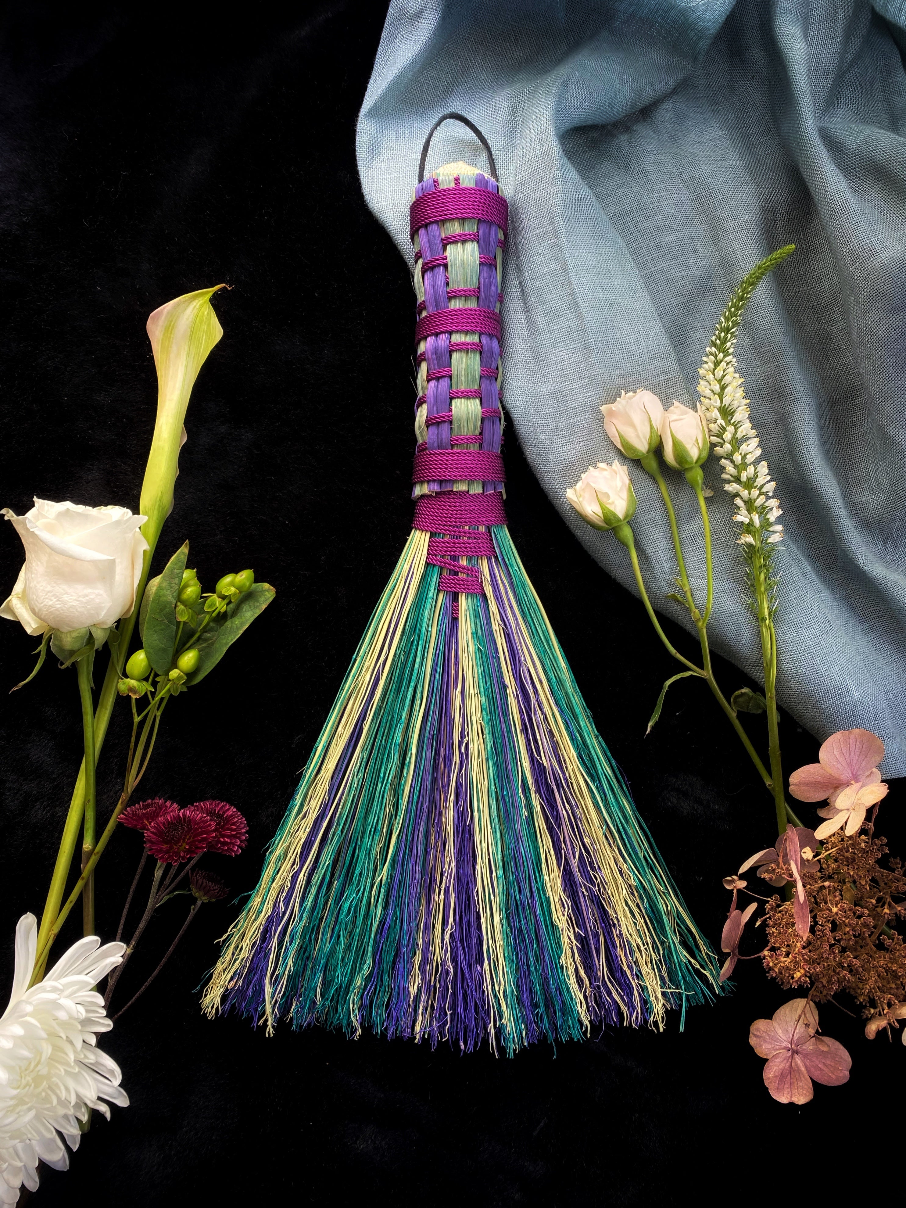 A hand-dyed, woven handle hawk tail broom with a multicolored handle and a green and beige bristle, displayed among various flowers against a black background.