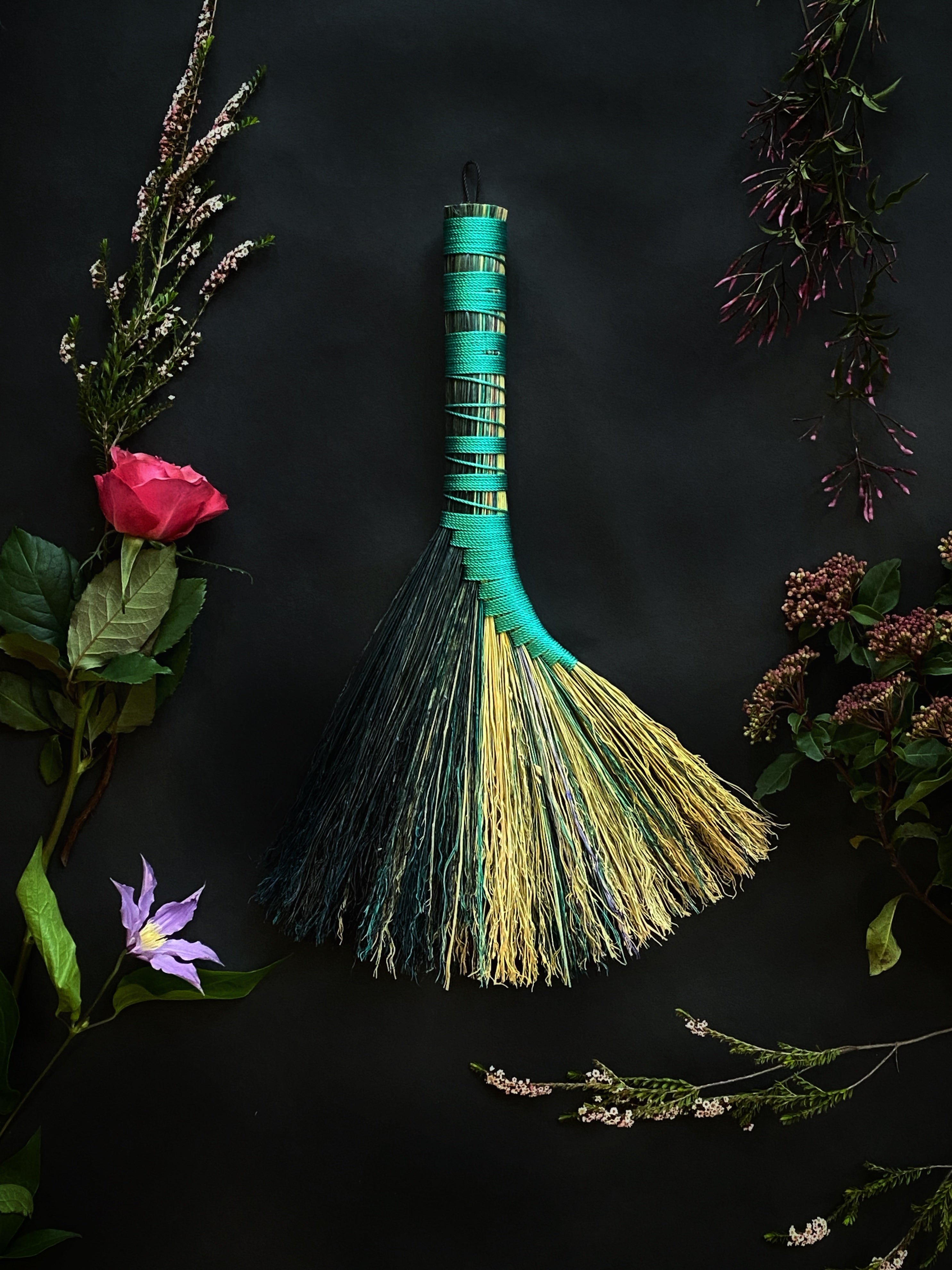 A handcrafted broom with a turquoise handle and multicolored bristles, displayed among various flowers and foliage.